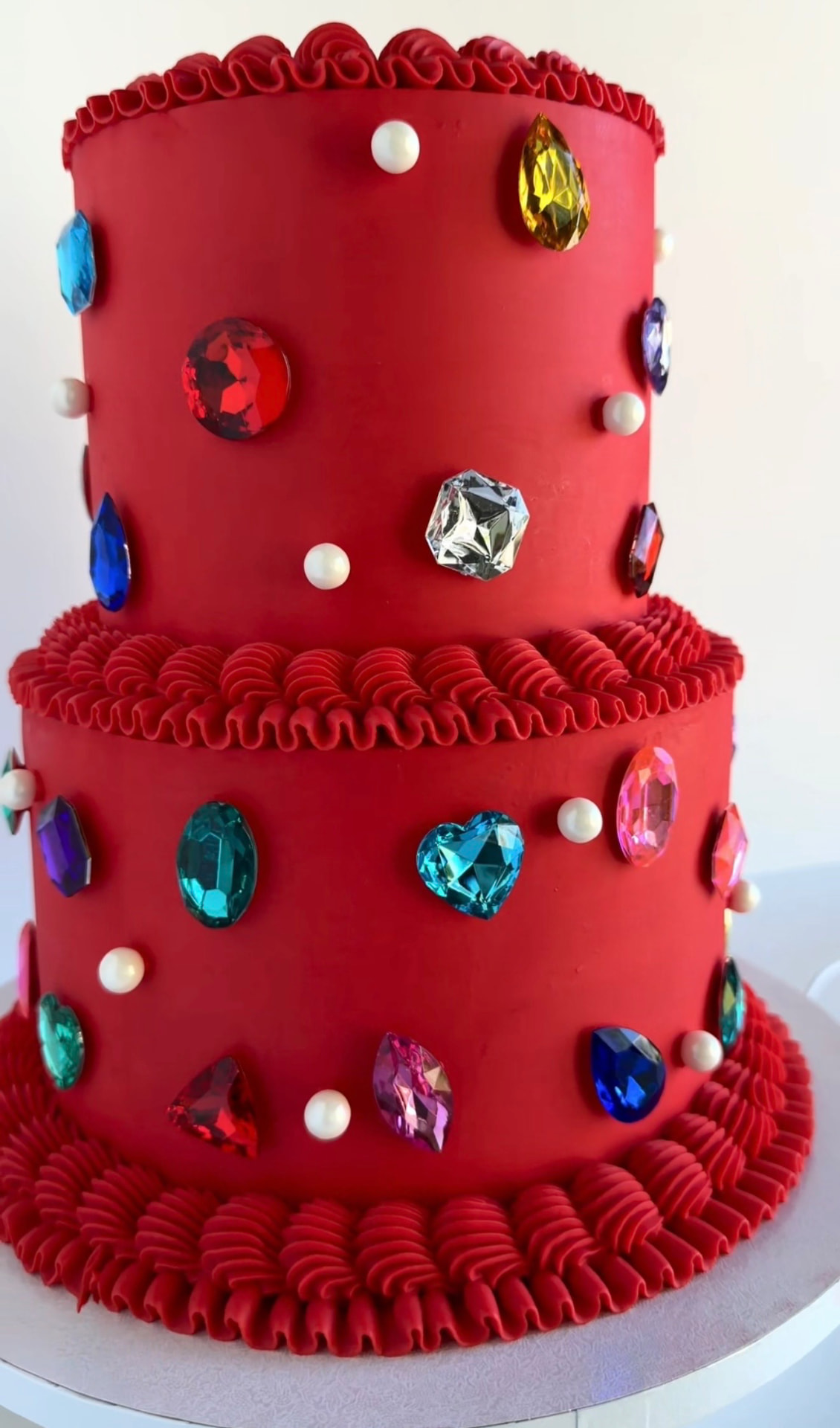 Two-tier red cake with piped ruffle borders, colorful gem accents and pearls