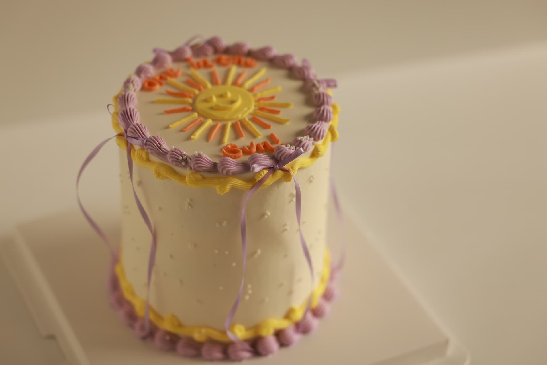 Cream cake with a piped sunshine face, purple scalloped swags and flowing lavender ribbons